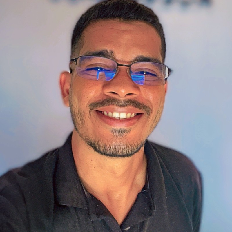 Foto de Paulo Glebson sorrindo, usando óculos e camisa preta, barba e fundo branco.